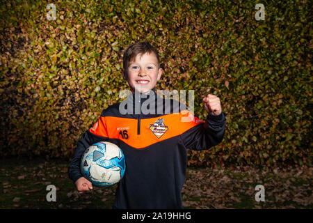 Ein acht Jahre alter Junge trägt eine Fußball-Streifen und halten ein Fußball unter dem Arm, mit der anderen Hand in eine Faust. Stockfoto