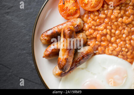Eine Mahlzeit mit gegrillten Würstchen, gegrillte Tomaten, zwei Spiegeleier und gebackene Bohnen. Dunkle Schiefer Hintergrund. Dorset England UK GB Stockfoto
