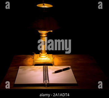 Moody, dunkles Bild eines Notebooks mit einem Bleistift auf einem Holztisch, beleuchtet von einer alten Vintage Lampe Stockfoto