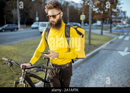 Junge Bartgeier Kurier liefern Essen auf einem Fahrrad, überprüfen Sie den mit einem Smartphone während in der Stadt stehen. Lieferung Service Konzept Stockfoto