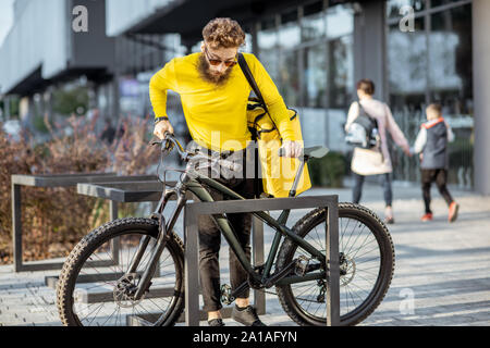 Junge Bartgeier Kurier Lieferung von Nahrung mit thermischen Beutel, Parkplatz sein Fahrrad auf der Straße in der Nähe des Büros. Lieferung Service Konzept Stockfoto