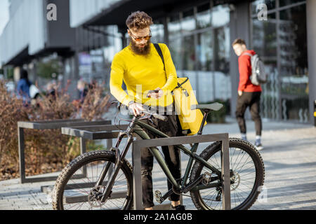 Junge Bartgeier Kurier Lieferung von Nahrung mit thermischen Beutel, Parkplatz sein Fahrrad auf der Straße in der Nähe des Büros. Lieferung Service Konzept Stockfoto