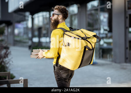 Junge männliche Kurier in Gelb sweatshirt liefern Pizza, stehend mit thermischen Rucksack auf dem steret im Freien Stockfoto