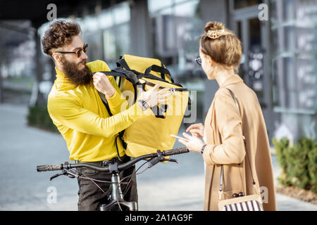 Männliche Kurier Lieferung von Nahrung mit einem Fahrrad in eine junge geschäftsfrau, bekommen einige Pakete aus einer thermischen Tasche im Freien Stockfoto