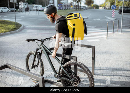 Junge Bartgeier Kurier Lieferung von Nahrung mit thermischen Beutel, Parkplatz sein Fahrrad auf der Straße in der Nähe des Büros. Lieferung Service Konzept Stockfoto