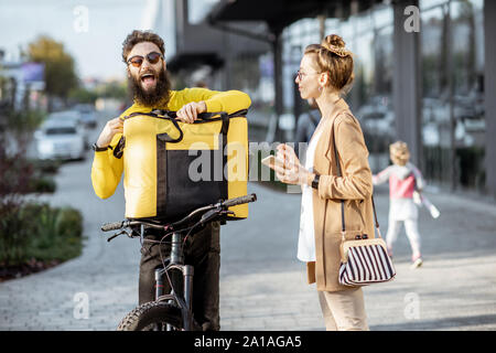 Männliche Kurier Lieferung von Nahrung mit einem Fahrrad in eine junge geschäftsfrau, bekommen einige Pakete aus einer thermischen Tasche im Freien Stockfoto