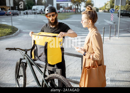 Männliche Kurier Lieferung von Nahrung mit einem Fahrrad in eine junge geschäftsfrau, bekommen einige Pakete aus einer thermischen Tasche im Freien Stockfoto