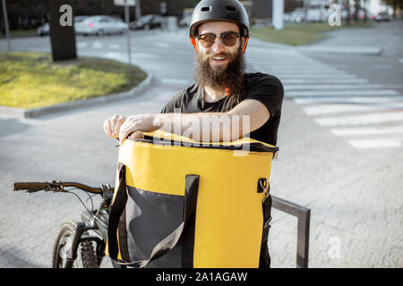 Portrait eines glücklichen jungen Kurier stehend mit Fahrrad und Gelb thermal Bag für Essen in der Stadt. Stockfoto