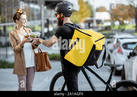 Kurier liefern frische Mittagsmenüs an eine junge geschäftsfrau auf einem Fahrrad mit Rucksack. Takeaway Restaurant essen Konzept Stockfoto