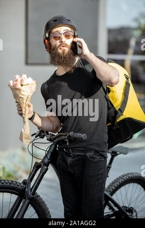 Jung und fröhlich Kurier warten mit Blumen für einen Kunden in der Nähe der Gebäude im Freien, frischen Floristik auf einem Fahrrad mit thermischen Tasche Stockfoto