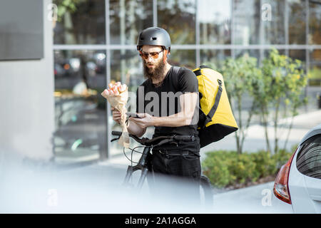 Jung und fröhlich Kurier warten mit Blumen für einen Kunden in der Nähe der Gebäude im Freien, frischen Floristik auf einem Fahrrad mit thermischen Tasche Stockfoto