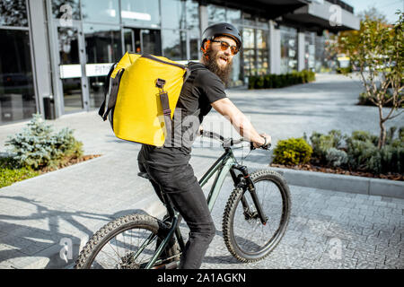 Junge Bartgeier Kurier Lieferung von Nahrung mit einem gelben thermische Rucksack, mit dem Fahrrad in der Stadt. Food Delivery Service Konzept Stockfoto
