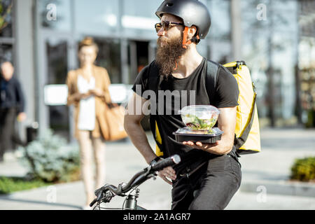 Kurier warten auf den Client mit Lunchpakete zum Mitnehmen, stehend auf einem Fahrrad in der Nähe des Bürogebäudes. Takeaway Restaurant essen Konzept Stockfoto