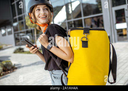 Portrait einer jungen und freundlichen weiblichen Kurier stehend mit Smart Phone und Gelb thermische Rucksack, Bereitstellung von frischen Produkten im Freien Stockfoto