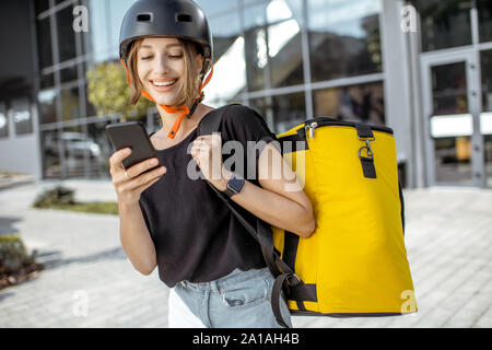 Portrait einer jungen und freundlichen weiblichen Kurier stehend mit Smart Phone und Gelb thermische Rucksack, Bereitstellung von frischen Produkten im Freien Stockfoto