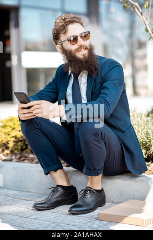 Porträt eines eleganten bärtigen Geschäftsmann im Anzug sitzen mit Telefon in der Nähe des Bürogebäudes outdooors gekleidet Stockfoto