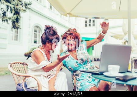 Zwei aufgeregt älterer Frauen lesen gute Nachrichten auf der Linie mit einem Laptop im Café im Freien. Stockfoto