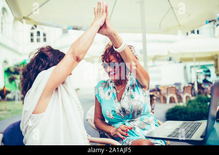 Zwei glückliche ältere weibliche Freunde geben High Five, Erfolg feiern, während Sie einander sitzen, die in der Tabelle in einem Café im Freien. Stockfoto