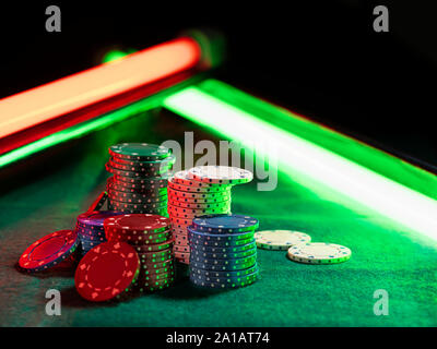 - Aus der Nähe geschossen von einem bunten chips Piles, einige von ihnen Festlegung in der Nähe auf grüne Abdeckung von Spielen Table unter grünen und roten Neonlichtern. Schwarz Stockfoto