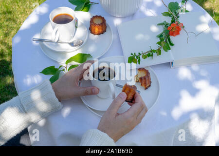 Genießen Kaffee Zeit im Garten. Serviert Tisch mit Blumen, Buch, Kaffee und Dessert im Garten im Frühling. Anordnung mit den Händen einer Frau. Stockfoto