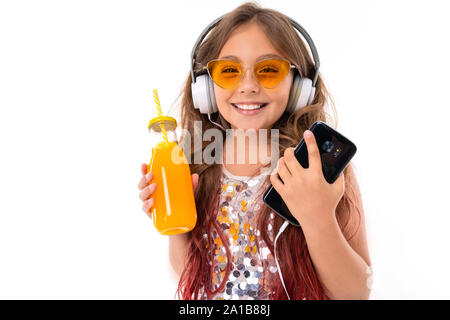 Schöne Mädchen im glitzernden Kleid und helle gelbe Sonnenbrille, mit großen weißen Ohrhörer, Musik hören und halten BlackBerry-Smartphone und Flasche mit Stockfoto