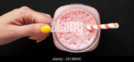 Weibliche hand mit einem Glas Glas mit Strawberry Banana Smoothie über schwarze Fläche gefüllt, Ansicht von oben. Ansicht von oben, von oben, flach. Nahaufnahme. Stockfoto