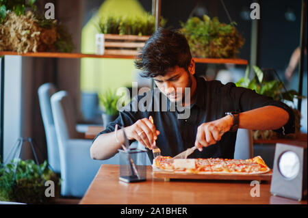 Selbstbewussten jungen indischen Mann in Schwarz Shirt in der Pizzeria mit Pizza sitzen. Stockfoto