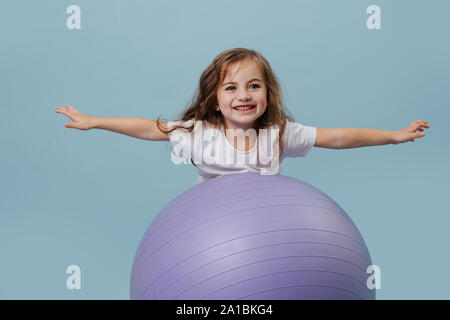 Kleines Mädchen versucht, auf eine Yoga Ball ohne Hände über blauen Hintergrund zu klettern Stockfoto