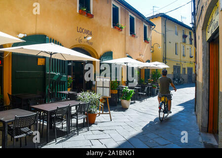 Ein Mann mit dem Fahrrad in eine enge Gasse des historischen Zentrums von Lucca mit einem leeren Restaurant im Freien an einem sonnigen Sommertag, Toskana, Italien Stockfoto