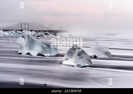 Eisblöcke auf einem schwarzen vulkanischen Strand entfernt sind, ist die Bewegung der Wellen gesehen werden kann (langzeitbelichtung), darüber ein bewölkter Himmel mit roten Schattierungen von Stockfoto