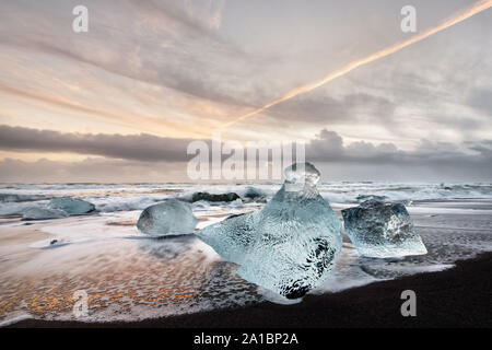 Eisblöcke in Blau und dunklen Schatten auf einem schwarzen Strand mit starker Brandung, das Eis in den nassen Sand reflektiert wird, Wellen im Hintergrund - Lage: I Stockfoto