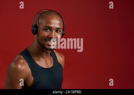 Fröhlicher Sportler. Portrait von gutaussehenden fröhlichen afro amerikanischen Mann in Kopfhörer Blick auf die Kamera, während vor rotem Hintergrund stehen. Sportkonzept. Aktiver Lebensstil Stockfoto