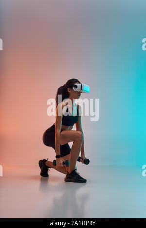 Die VR-Technologie. Seitenansicht der junge sportliche Frau in Sport Kleidung Training mit Hanteln beim Tragen von virtual reality Brillen. Studio shot Stockfoto