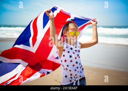 Gerne britische Mädchen trägt flattern Blau Weiß Rot Flagge von Großbritannien, des Vereinigten Königreichs und Nordirland gegen den blauen Himmel und das Meer zurück Stockfoto