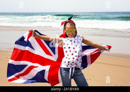 Gerne britische Mädchen trägt flattern Blau Weiß Rot Flagge von Großbritannien, des Vereinigten Königreichs und Nordirland gegen den blauen Himmel und das Meer zurück Stockfoto