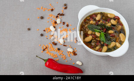 Mexikanische Suppe der sieben Arten von Bohnen mit Basilikum, Nahaufnahme, auf grauem Leinen Hintergrund von Paprika und Bohnen umgeben Stockfoto