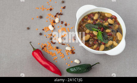Mexikanische Suppe der sieben Arten von Bohnen, Nahaufnahme, auf grauem Leinen Hintergrund durch rote und grüne Paprika und Bohnen umgeben