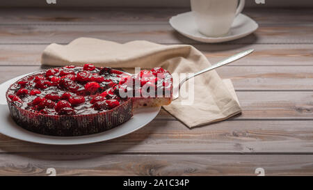 Torte mit Beeren: Himbeeren, Erdbeeren, Johannisbeeren, auf einem weissen Teller, neben einem Stück Kuchen auf der Schulter. Auf einer hölzernen Hintergrund, in der Stockfoto