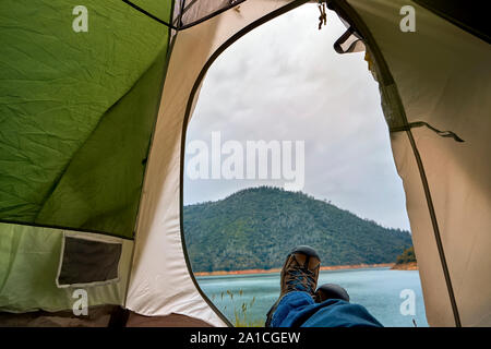 Wanderschuhe an den Eingang eines Zeltes mit einem Ausblick auf die Berge und den See unter einem bewölkten Himmel. Stockfoto