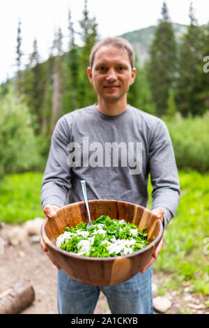 Junge Mann hält kale Salat große hölzerne Schüssel mit Hintergrund der grünen Utah Berge in Albion Basin Campground Stockfoto