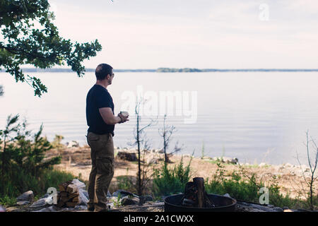 Mitte Alter kaukasischer Mann steht an der campside durch den See bei einer Tasse Kaffee Stockfoto