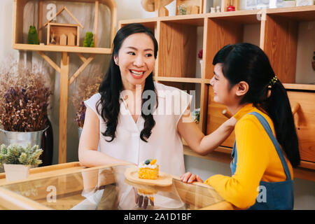 Lieben asiatischen Mutter und Tochter im casual Outfit sharing Gebäck am Tisch sitzen und an jedem anderen Suchen Stockfoto