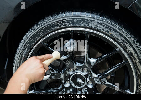 Ein männlicher Arbeitnehmer wäscht ein schwarzes Auto mit einer speziellen Bürste für Gegossene Räder und scheuert sich die Oberfläche zu glänzen in einem Fahrzeug mit Werkstatt. Auto Service in Stockfoto