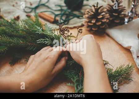 Hände, die Kräuter und Tannenzweigen, Tannenzapfen, Gewinde, Beeren, Zimt auf Holztisch. Weihnachten Kranz Werkstatt. Authentische stilvolle noch leben. Stockfoto