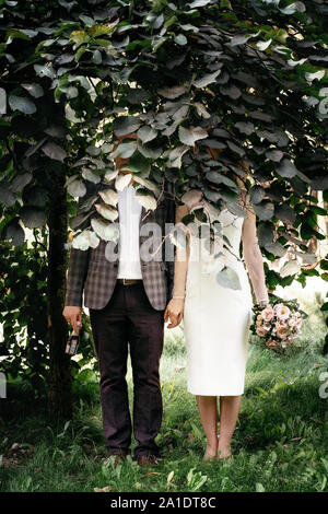 Hochzeit paar steht unter grünen Bäumen mit Flächen von Laub und halten die Hände versteckt Stockfoto