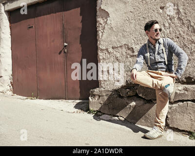 Junge Mann in Strapsen und Sonnenbrille in der Sonne zu sitzen vor einem Haus im ländlichen Raum Stockfoto