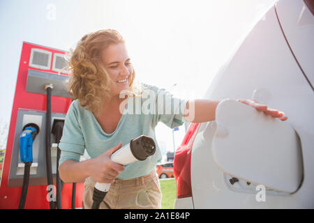 Junge Frau steht in der Nähe des Elektroautos. Das Auto wird der Ladevorgang an der Ladestation für Elektrofahrzeuge. Stockfoto