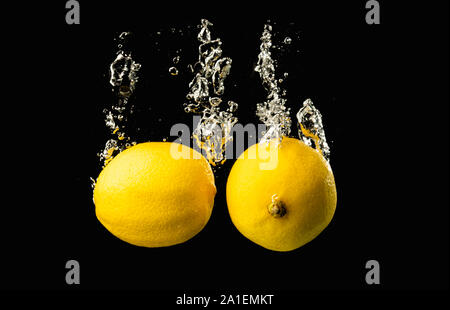 Frische gelbe Zitronen in Wasser spritzen auf schwarzen Hintergrund mit vielen Luftblasen. Stockfoto