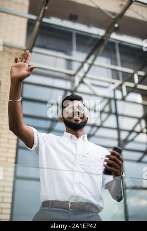 Ernsthafte indischer Geschäftsmann in Brillen suchen auf Kopieren, während sich gegen Glashochhaus winken Hallo zu empfehlen. Stockfoto