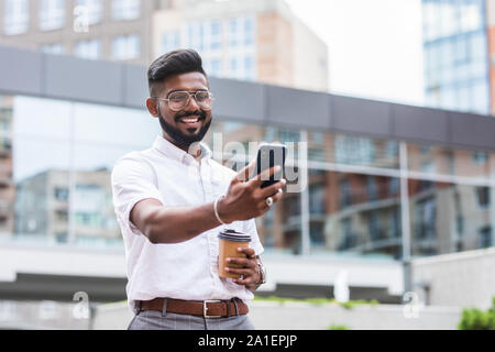 Indischer Geschäftsmann ist entspannend in der Stadt und winken in die Kamera von seinem Telefon in einer Videokonferenz Stockfoto
